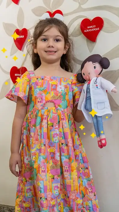 Banner Dona Rosinha — boneca educativa da Alphabeto. Menina sorrindo segurando a boneca Dona Rosinha, em frente a uma árvore decorativa com corações vermelhos com nomes e o logo Alphabeto.
