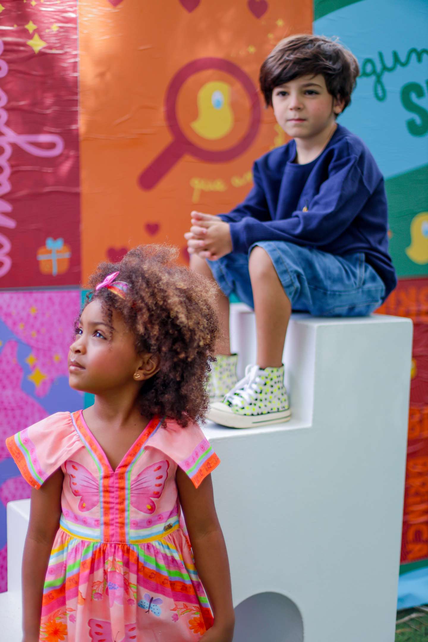 A imagem mostra uma menina sorridente em frente a um fundo com formas geométricas coloridas e a frase "Oba, tem lançamento no nosso mundo mágico". 