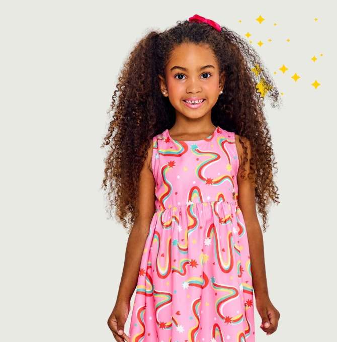 A imagem mostra uma menina sorridente com cabelos soltos em frente a um fundo neutro com estrelinhas amarelas. A enina veste um vestido rosa estampado. 