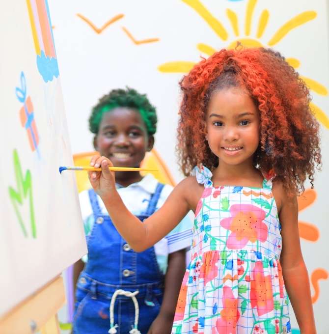 A imagem mostra duas meninas sorridentes posando em frente a um fundo laranja vibrante com um grande círculo texturizado rosa ao centro. A menina à esquerda usa uma roupa colorida com estampas divertidas, jaqueta clara e tiara. A menina à direita veste blusa preta e saia rosa. O ambiente é alegre e lúdico.
