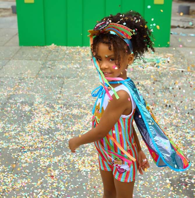 A imagem mostra uma menina vestida com uma fantasia da Alphabeto em meio a uma chuva de confetes de carnavall