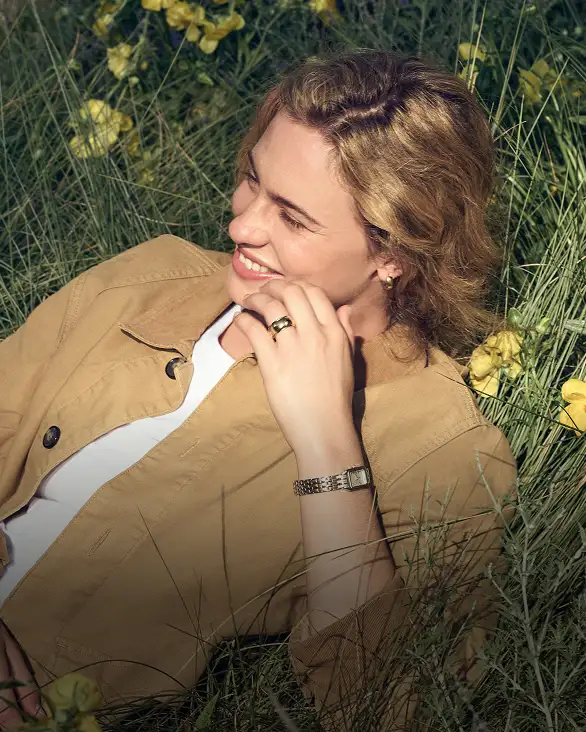 A woman wearing a Fossil watch and jewelry, lounging in a spring meadow among yellow flowers.