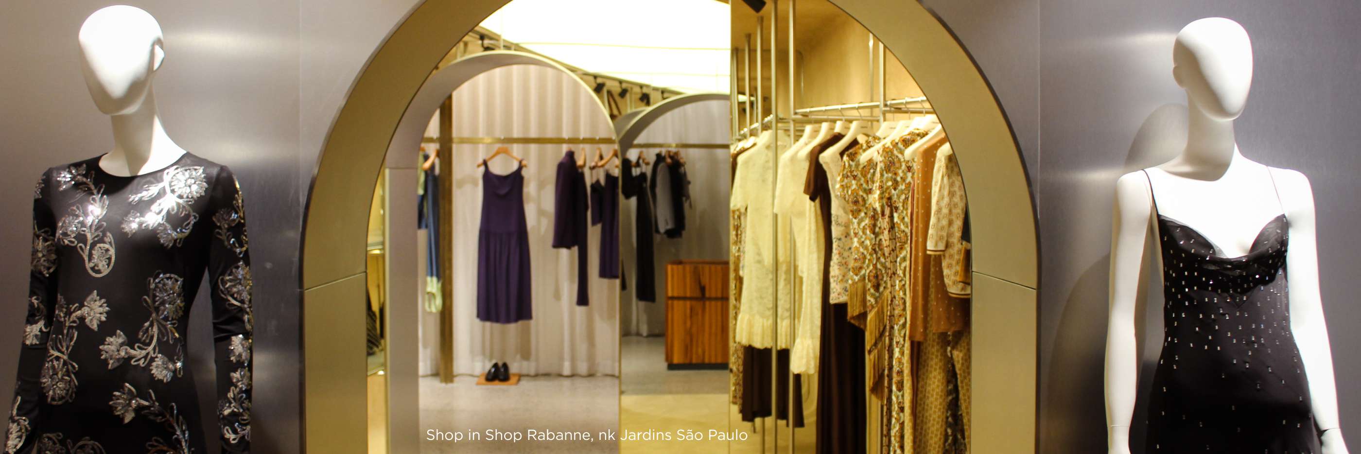 A imagem captura uma boutique da marca Rabanne com um design arquitetônico deslumbrante, caracterizado por arcos dourados e elegantes que emolduram diferentes seções da loja. As roupas, penduradas em cabides metálicos, apresentam uma paleta de cores suaves com vestidos em tons de roxo, bege, dourado e estampas delicadas, refletindo o estilo sofisticado e contemporâneo da marca. O ambiente, localizado no shopping Jardins em São Paulo, combina elementos arquitetônicos modernos com uma iluminação suave que destaca as peças, criando um espaço de varejo que é tanto uma galeria de moda quanto um ambiente de compras convidativo e luxuoso. 