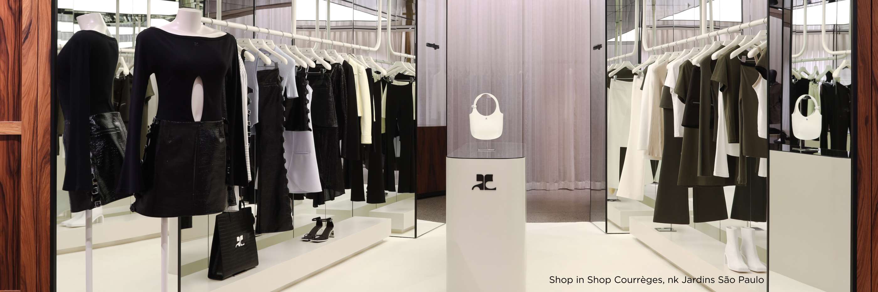 A imagem retrata uma boutique da marca Courrèges com design minimalista, apresentando uma coleção elegante em tons de preto, branco e verde militar. Os manequins exibem peças de vestuário contemporâneas penduradas em cabides brancos, dispostos em um ambiente ampliado por espelhos e iluminação suave. No centro, destaca-se um pedestal branco com uma bolsa circular e sapatos pretos, criando um cenário sofisticado que reflete o estilo refinado da marca. Localizada no shopping Jardins em São Paulo, a loja transmite uma estética moderna e clean, convidando os clientes a experimentar uma moda minimalista e atual.