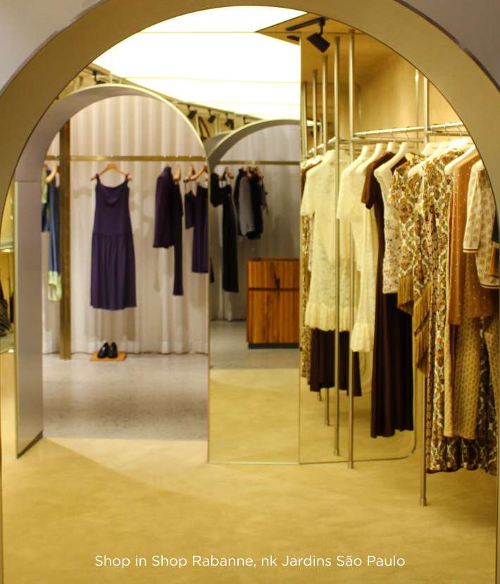 A imagem captura uma boutique da marca Rabanne com um design arquitetônico deslumbrante, caracterizado por arcos dourados e elegantes que emolduram diferentes seções da loja. As roupas, penduradas em cabides metálicos, apresentam uma paleta de cores suaves com vestidos em tons de roxo, bege, dourado e estampas delicadas, refletindo o estilo sofisticado e contemporâneo da marca. O ambiente, localizado no shopping Jardins em São Paulo, combina elementos arquitetônicos modernos com uma iluminação suave que destaca as peças, criando um espaço de varejo que é tanto uma galeria de moda quanto um ambiente de compras convidativo e luxuoso. 